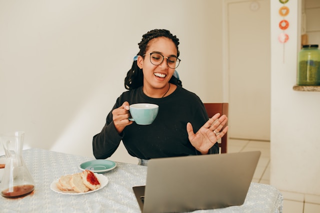 Woman wearing black glasses, holding a blue coffee cup, and waving at a computer monitor in front of her.