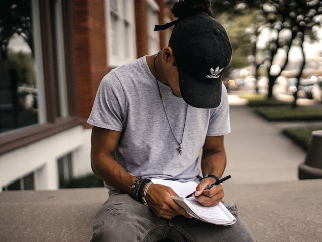 Man sitting on a brick wall writing in a notebook.‌