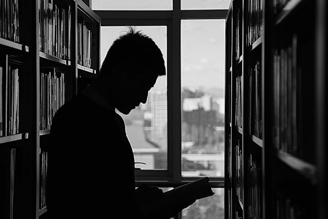 Silhouette of a man reading a book between bookshelves.