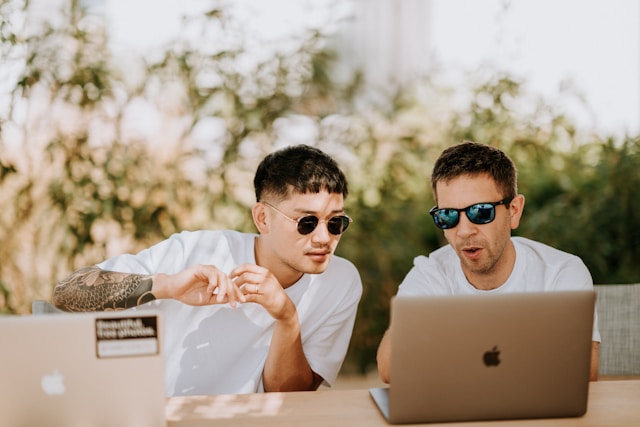 Two men wearing sunglassases sitting at a table next to each other and looking at one person’s laptop screen.