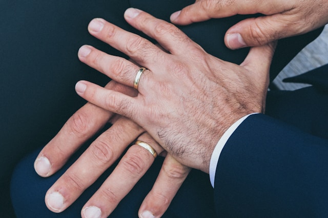 08-14-24b_nick-karvounis-DUK7XiBpGSY-unsplash Two male-presenting people’s left hands atop each other with both wearing gold wedding rings.