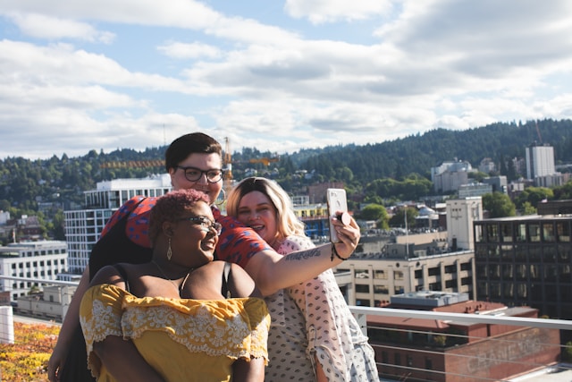 A trio of plus-size women with broad smiles posing for a selfie on a city roof in the daytime.