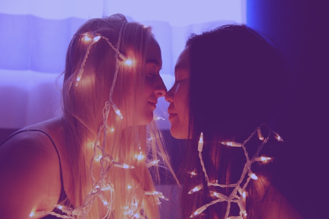 Sweet romance or burning-hot spice, queer romance readers deserve a story that will make them wish for their own perfect partner. A blonde woman and black-haired woman nose-to-nose and smiling, with white Christmas lights draped around them.
