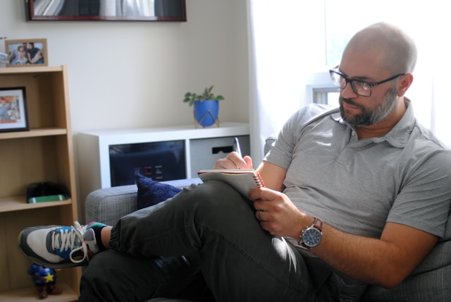 At a certain point, I accept that the choices my characters make are no longer up to me. Bald man wearing black glasses, dressed in a collared gray shirt, black pants, and tennis shoes, writing in a notepad.