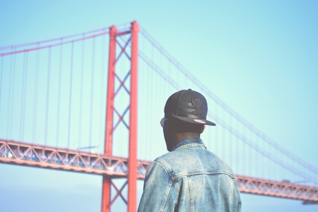 Transition scenes are important connection points between the major events of your story. Man in black hat and light blue jean jacket with back to camera, looking toward red suspension bridge.