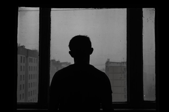 Experiencing heartache in real life is devastating, no matter how it shows itself. Silhouette of a man standing in front of a window looking out at an urban area.