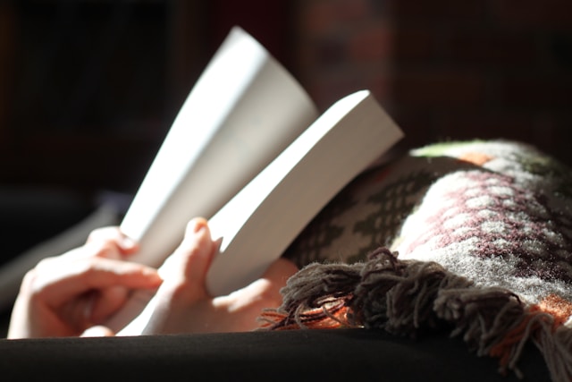 Reading a book and writing a book are two sides of the creative journey coin. Closeup of a person under a patterned blanked reading a paperback book.